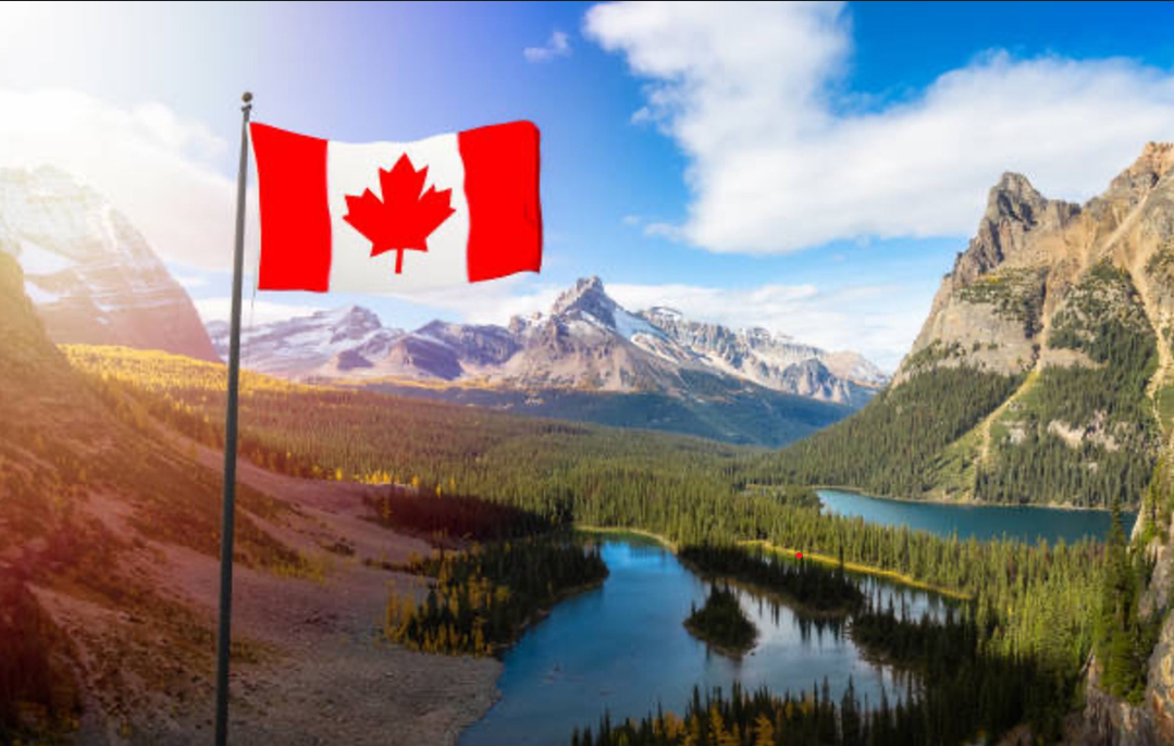 A Canadian flag waving on a flagpole overlooking a picturesque mountain valley with a clear blue lake, green pine forests, and rocky peaks under a sunny sky.
