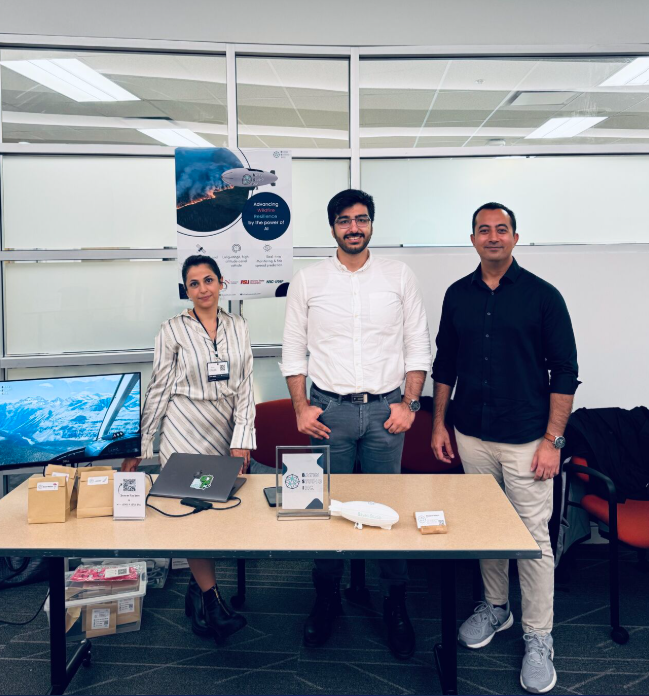 Three Bayes Studio team members standing behind a display table with a laptop, branded display signs, and a miniature airship model at Entrepreneurship@UBC Immersion Week 2024.