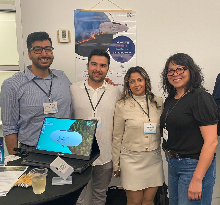 Four Bayes Studio team members standing beside a display table at the Innovation UBC Investor Showcase, featuring a monitor showing a high-altitude aerial blimp rendering and an official research poster titled "Advancing Wildfire Resilience by the power of AI."