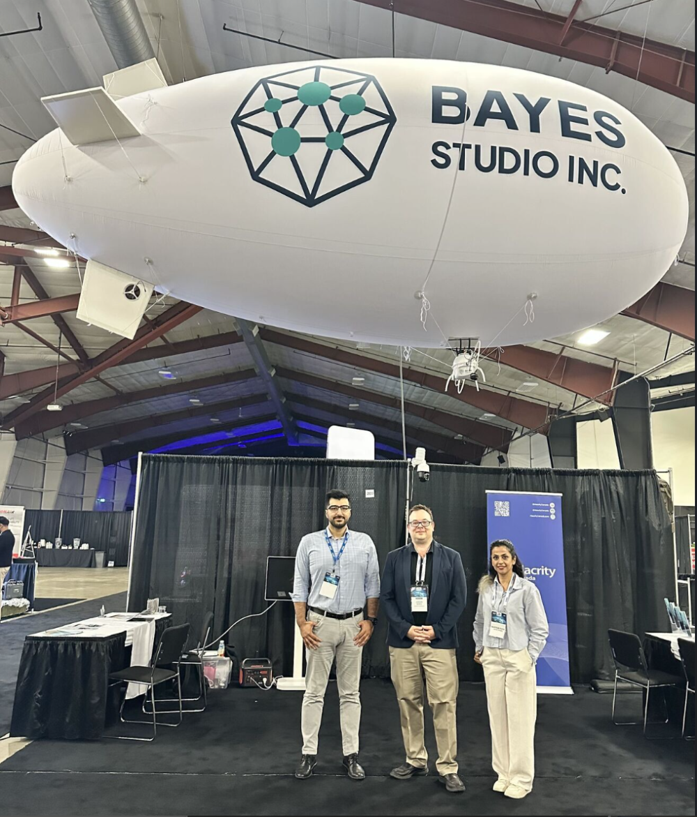 A photograph taken at the Bayes Studio booth at the ADSE 2026 expo. Three team members stand smiling in front of a black curtain backdrop. Hovering above them is the large, white, blimp-style autonomous UAV called 'Helius O'. The UAV features the 'Bayes Studio Inc.' text logo and their teal and blue geometric icon clearly displayed on the side.