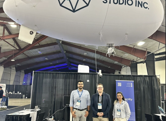 A photograph taken at the Bayes Studio booth at the ADSE 2026 expo. Three team members stand smiling in front of a black curtain backdrop. Hovering above them is the large, white, blimp-style autonomous UAV called 'Helius O'. The UAV features the 'Bayes Studio Inc.' text logo and their teal and blue geometric icon clearly displayed on the side.
